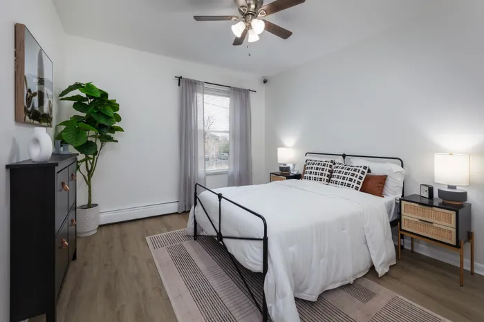 Spacious bedroom with ceiling fan, large window, vinyl wood flooring, decorated with white bedding, grey curtains and rug under the bed.