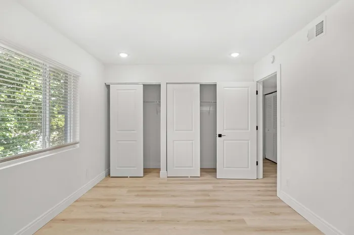 Bright and spacious bedroom with large window, showcasing natural light, sleek wood flooring, and ample closet storage.