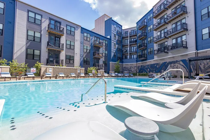 Modern apartment complex with vibrant blue accents and a luxurious pool area complete with stylish lounge chairs and ample sunbathing space.
