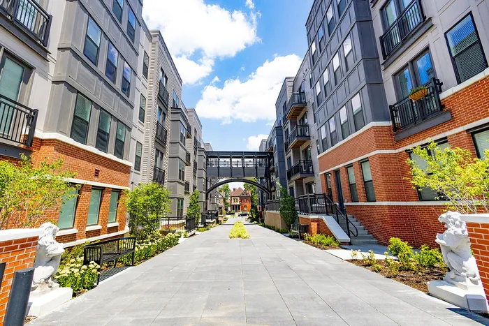 Modern apartment complex with stylish brick and gray facades, featuring landscaped pathways and a charming sky bridge.