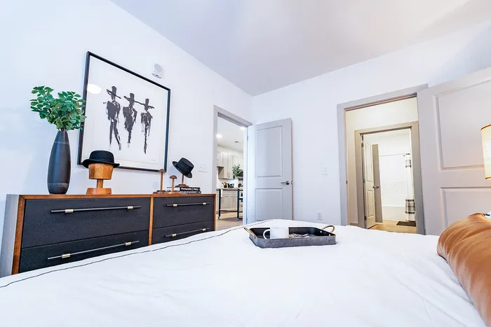 Bright and modern bedroom with sleek decor, stylish dresser, and open doorway to kitchen and bathroom.