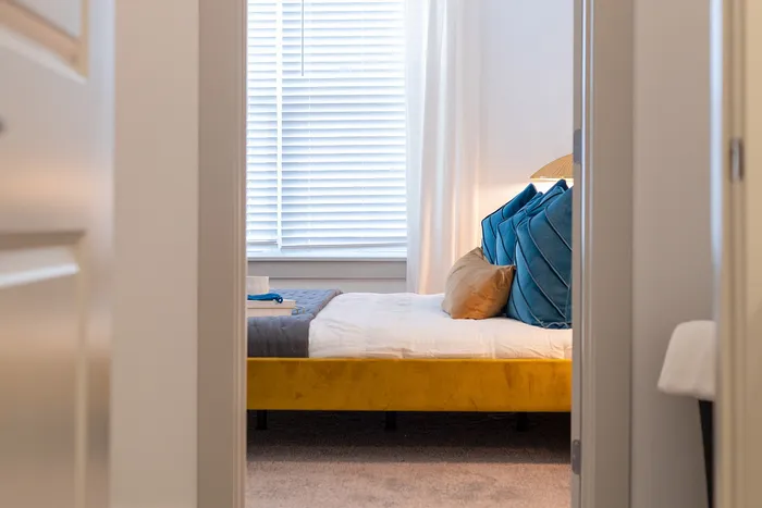 Inviting bedroom with vibrant yellow bed frame and plush blue pillows, bathed in natural light from large windows with blinds.