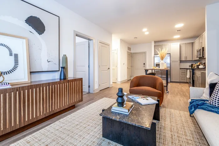 Modern open-concept living room with stylish decor, featuring a cozy brown chair, elegant artwork, and a sleek kitchen in the background.