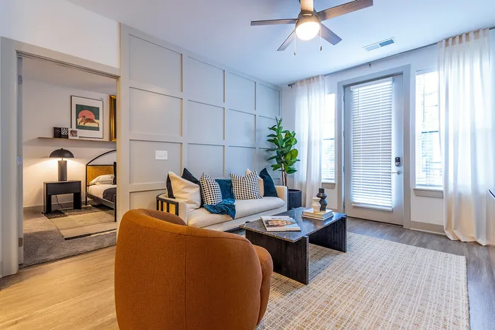 Inviting living room with modern decor, featuring a cozy beige sofa, vibrant cushions, and an elegant coffee table. Bright natural light fills the space through large windows with sheer curtains.