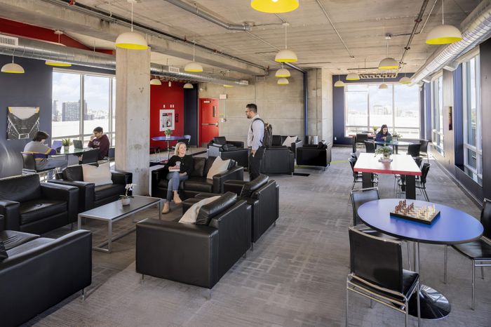 Modern communal lounge with chic black sofas, natural light from large windows, vibrant red accents, and people enjoying a relaxed atmosphere.