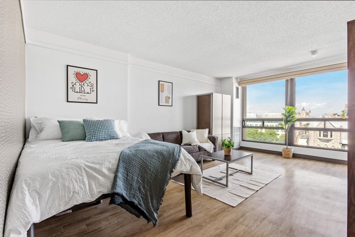 Chic studio apartment with a cozy bed, stylish sitting area, and expansive city views through large windows.