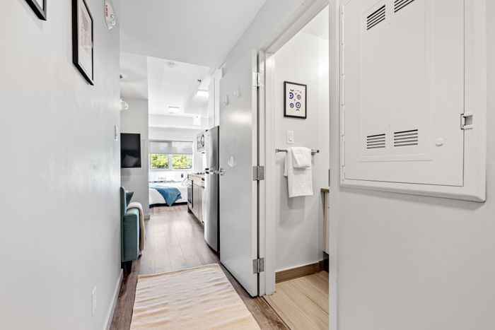Bright and modern apartment hallway leading to a cozy bedroom with large windows, featuring sleek appliances and elegant decor.