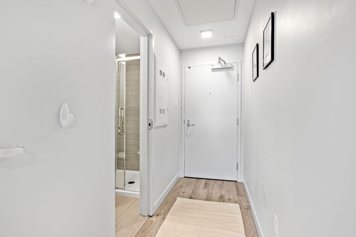 Bright apartment hallway with modern wood flooring, leading to a sleek bathroom with glass shower doors.