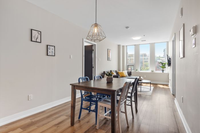 Bright and inviting apartment dining area with a modern wooden table, colorful chairs, and sunlit living space featuring large windows and stylish decor.