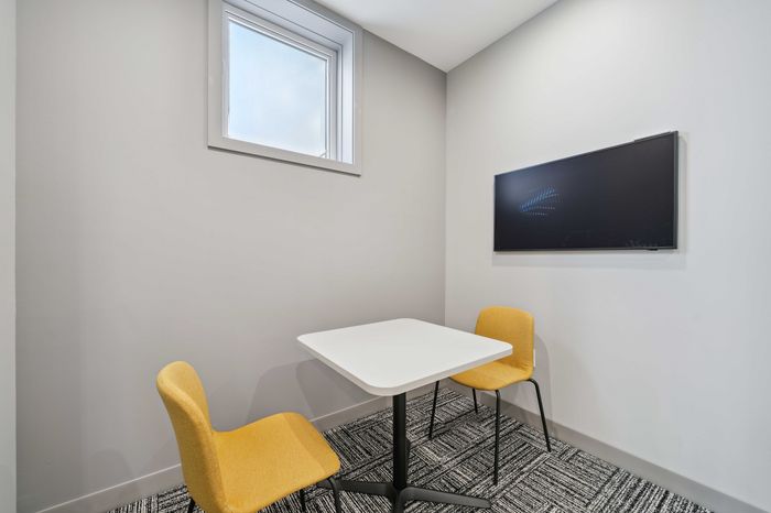 Sleek modern meeting nook with a minimalist table, vibrant yellow chairs, natural light from a window, and a sleek wall-mounted screen.