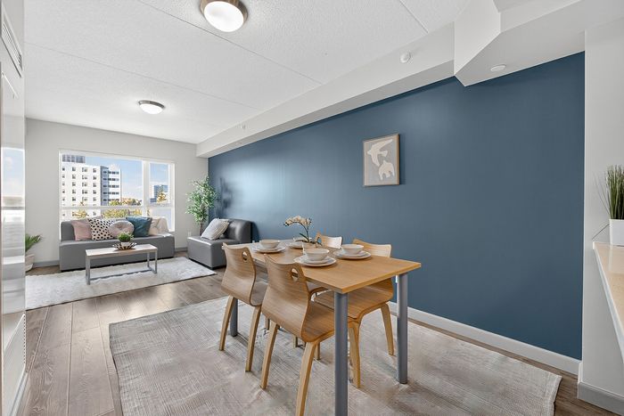 Modern apartment living area with a cozy dining table, stylish gray sofas, and a vibrant blue accent wall, filled with natural light from large windows.