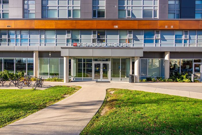 Modern apartment building entrance with large glass doors, lush green surroundings, and convenient bike racks, showcasing urban elegance and accessibility.
