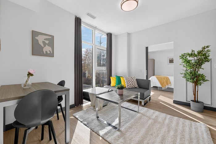 Bright and modern open-concept living area with large windows, featuring a cozy sitting space with colorful cushions, a small dining table with chic chairs, and a lush potted plant, leading into a bedroom with warm accents.