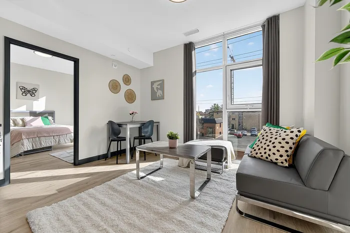 Bright and modern living space featuring a cozy gray sofa, stylish decor, large window with city view, and an inviting glimpse into a serene bedroom.