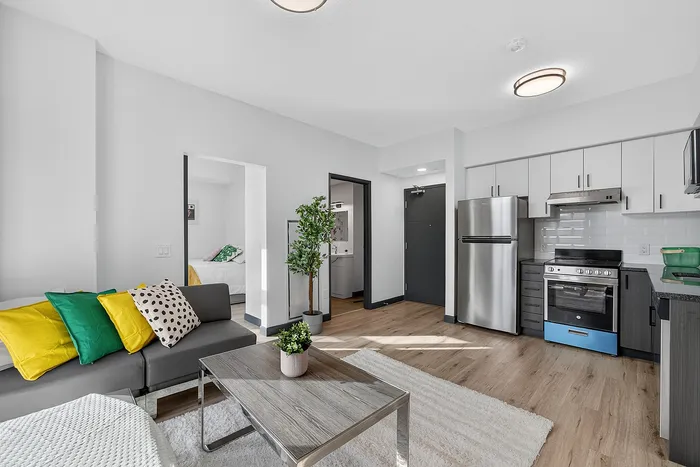 Modern open-plan apartment living room with stylish gray sofa, vibrant pillows, wooden coffee table, sleek kitchen with stainless steel appliances, and cozy bedroom entrance.