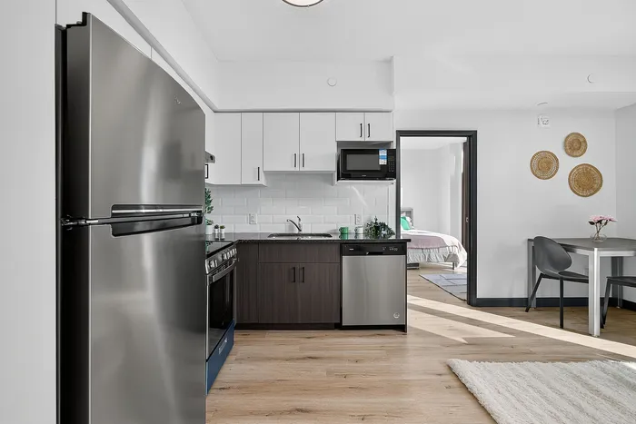 Contemporary apartment kitchen with sleek stainless steel appliances, elegant cabinetry, and cozy dining area, leading to a bright, inviting bedroom.