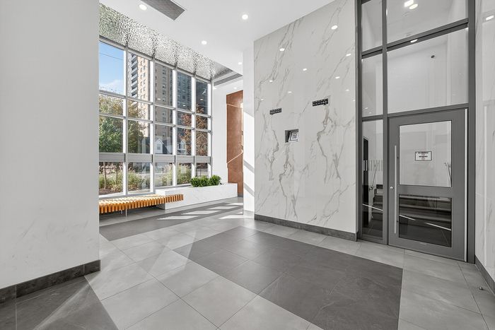 Bright and modern apartment lobby featuring floor-to-ceiling windows, elegant marble walls, and sleek tile flooring.
