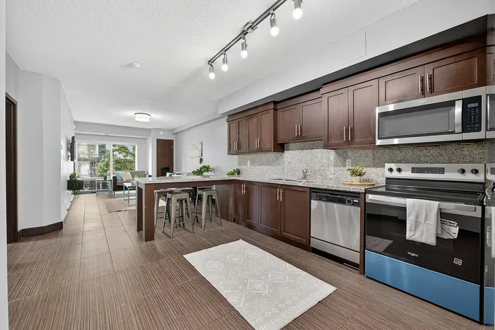 Modern kitchen and dining area with sleek dark wood cabinetry, stainless steel appliances, and bright track lighting leading to a cozy living room with garden views.