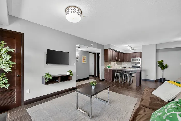 Modern living room with cozy seating, sleek coffee table, and wall-mounted TV, seamlessly connected to an open kitchen with stylish dark cabinetry and a breakfast bar.