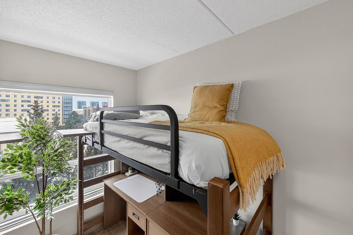 Bright apartment bedroom with a stylish loft bed, cozy yellow throw, and a desk beneath, offering scenic urban views.
