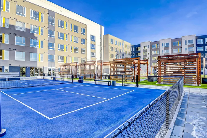 Modern apartment complex with vibrant tennis court and stylish wooden pergolas under clear blue skies.