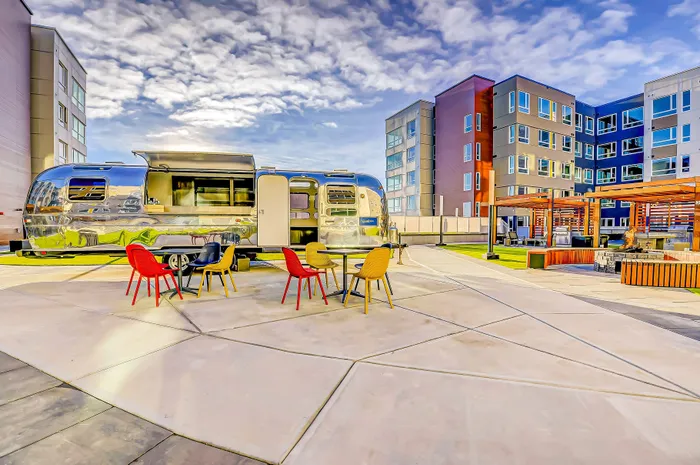 Vibrant urban patio featuring colorful chairs, a sleek Airstream trailer, and modern apartment buildings under a stunning sky.