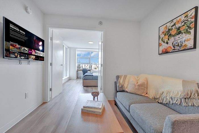 Bright, inviting apartment featuring a cozy living room with plush sofa, wall-mounted TV, and sunny bedroom through open doorway, offering modern comfort and style.