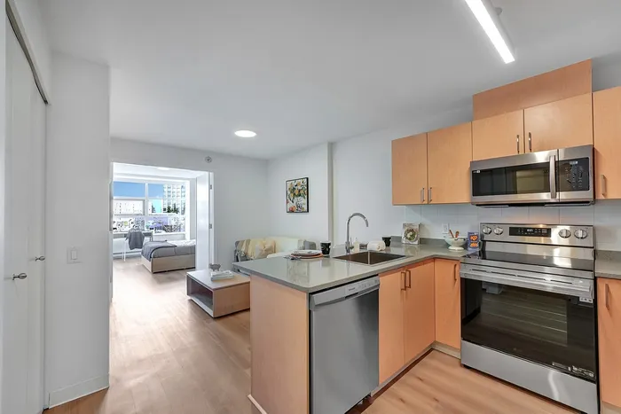 Modern kitchen with stainless steel appliances, light wood cabinets, and an open view leading to a bright bedroom in a stylish apartment.
