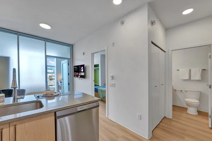 Modern open-concept kitchen and hallway featuring sleek countertops, stainless steel appliances, and light wood flooring, leading to a cozy bedroom and stylish bathroom.