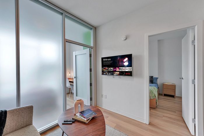 Modern apartment living area with stylish frosted glass partitions, a cozy workspace, and a wall-mounted TV. The inviting bedroom with natural light enhances the welcoming atmosphere.