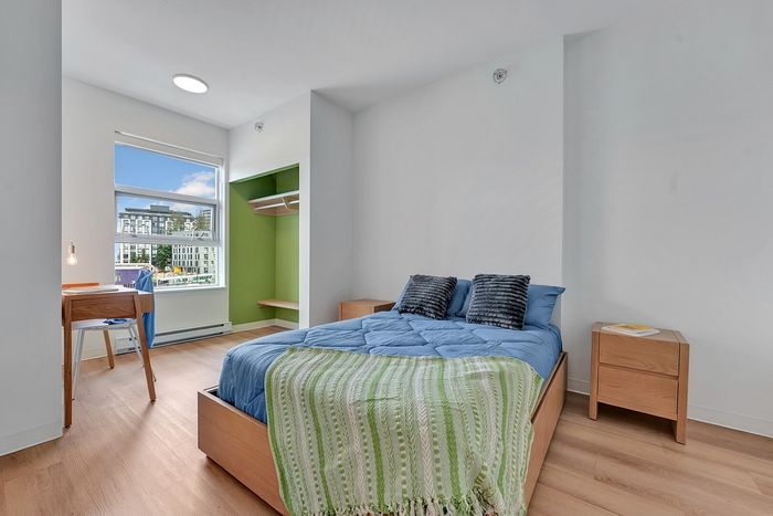 Bright, modern bedroom featuring a cozy bed with blue bedding, stylish wooden furniture, and a large window offering a scenic city view.