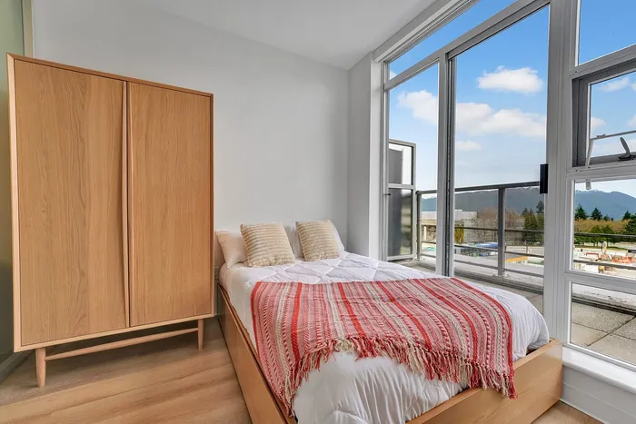 Bright bedroom featuring a cozy bed with a colorful blanket, large wooden wardrobe, and floor-to-ceiling windows with scenic views.