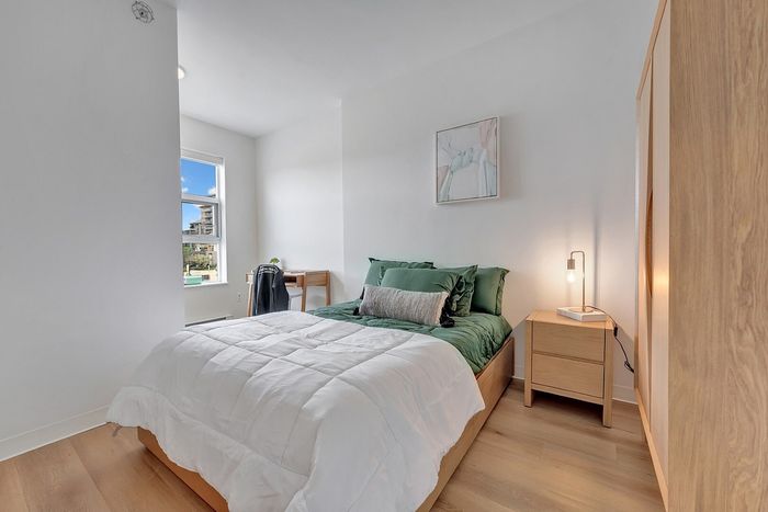 Bright and cozy bedroom with modern decor, featuring a comfortable bed with green accents, a wooden nightstand, and ample natural light from a large window.