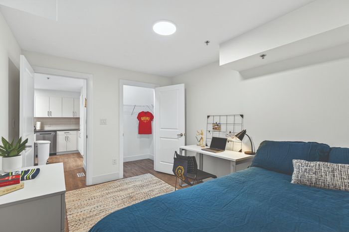 Modern bedroom with sleek workspace and closet, adjacent to a stylish kitchen with hardwood flooring and contemporary cabinetry.