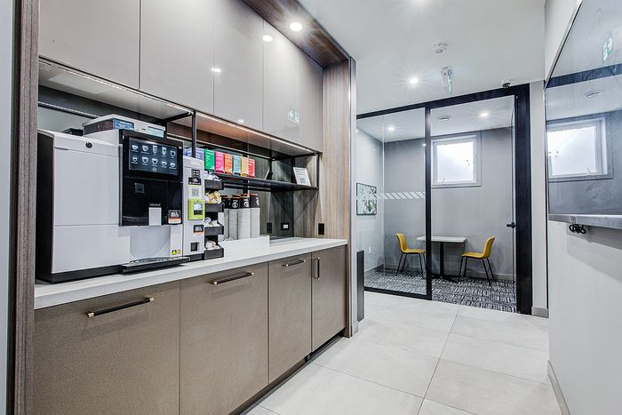 Modern kitchenette with a stylish coffee machine and sleek cabinetry, adjacent to a cozy meeting room with yellow chairs and a glass door.