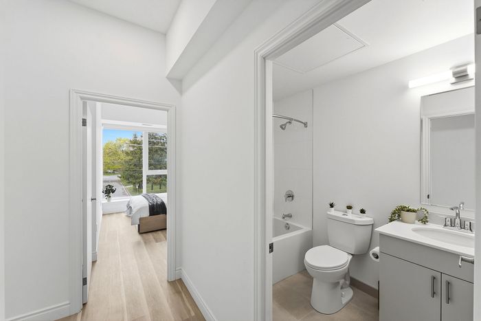 Bright apartment hallway with elegant hardwood flooring, leading to a cozy bedroom with large windows and a modern bathroom featuring stylish fixtures.