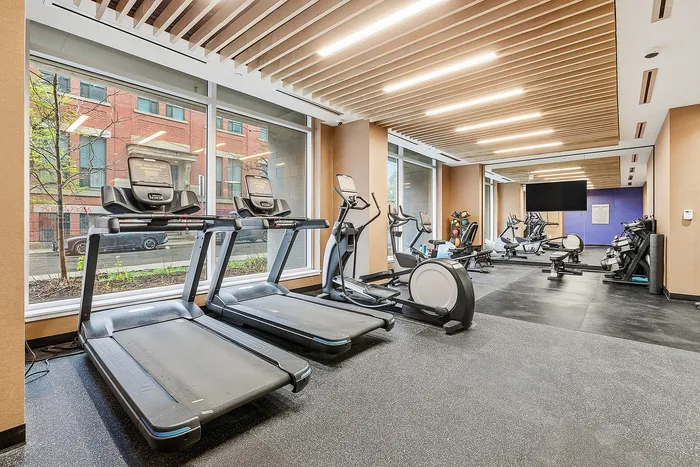 Bright and modern fitness center with state-of-the-art treadmills, elliptical machines, and a variety of workout equipment, set in a spacious area with large windows and stylish ceiling design.