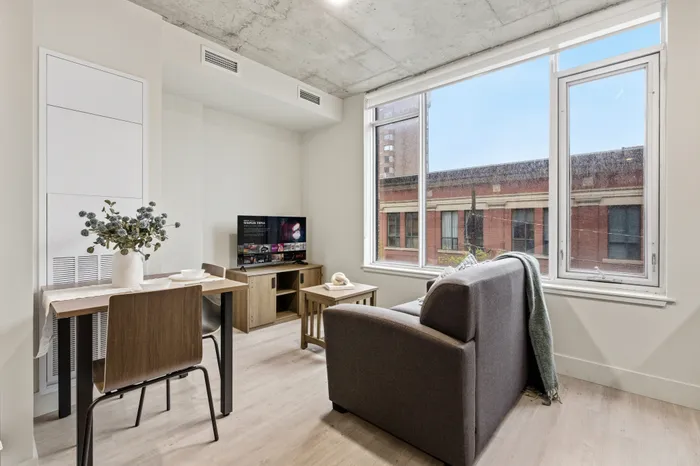Modern apartment living room with a cozy gray sofa, large window with city view, stylish dining area, and a TV setup.