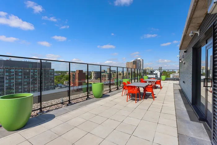 Vibrant rooftop terrace with modern red seating, expansive views of the city skyline, and lush green planters under a bright blue sky.