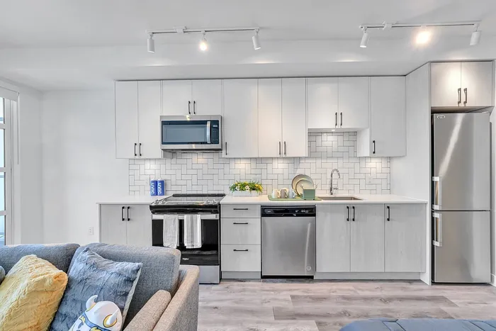 Modern kitchen with sleek white cabinetry, stylish backsplash, and stainless steel appliances, complemented by cozy living area seating.