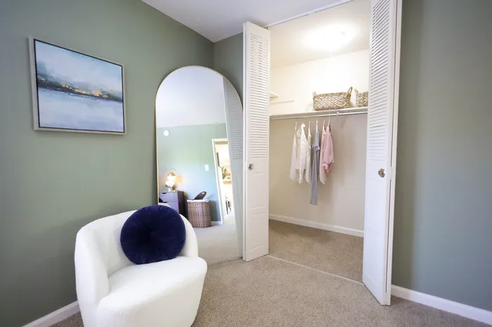 Elegant bedroom corner featuring a chic white armchair with a navy cushion, open closet for seamless storage, and soft green walls adorned with modern art.