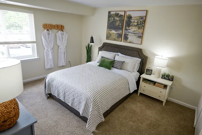 Inviting bedroom with a cozy bed, chic decor, and natural light from a large window. Features elegant wall art, soft carpeting, and a bedside table with stylish accents.