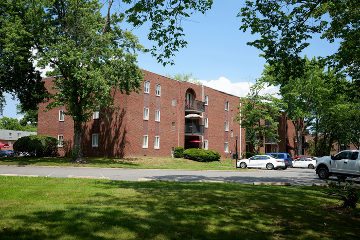 Charming brick apartment complex surrounded by lush green trees, offering relaxed suburban living with convenient parking.