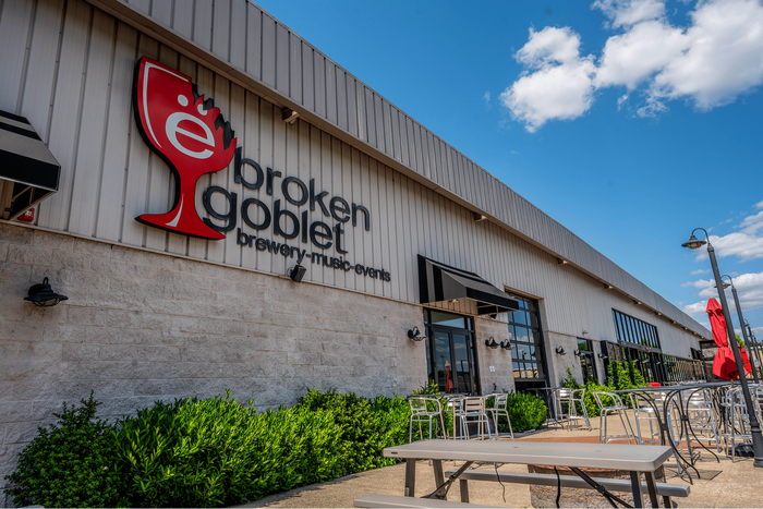 Modern brewery exterior with sleek design, featuring outdoor seating and vibrant red umbrellas under a clear blue sky.