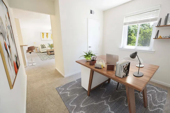 Bright home office with wooden desk, stylish decor, and large window overlooking green scenery, leading to an inviting open-concept living area with modern furnishings.