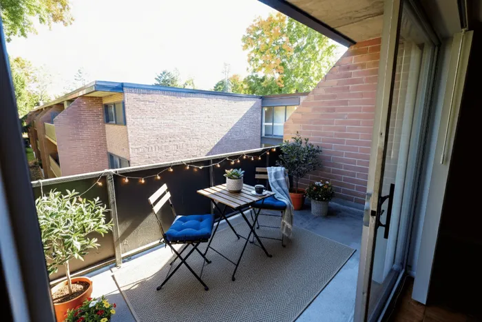 Charming apartment balcony with cozy seating, lush potted plants, and warm string lights under a sunny sky.
