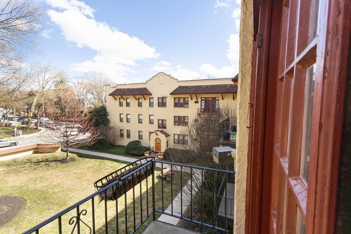Charming view from a balcony overlooking a classic brick apartment building with manicured lawn, surrounded by trees and blue sky.