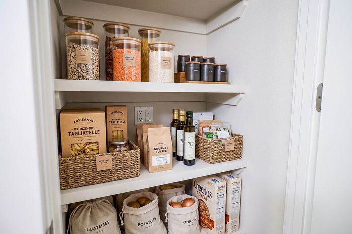 Immaculate pantry with organized shelves featuring glass containers of grains, artisanal pasta boxes, gourmet coffee, olive oil, and neatly arranged cereal boxes.