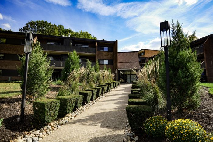 Inviting apartment exterior with a charming landscaped pathway lined with manicured hedges and vibrant greenery under a bright blue sky.