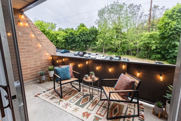 Charming balcony adorned with string lights, featuring cozy seating and vibrant pillows, overlooking lush greenery and a peaceful parking area.